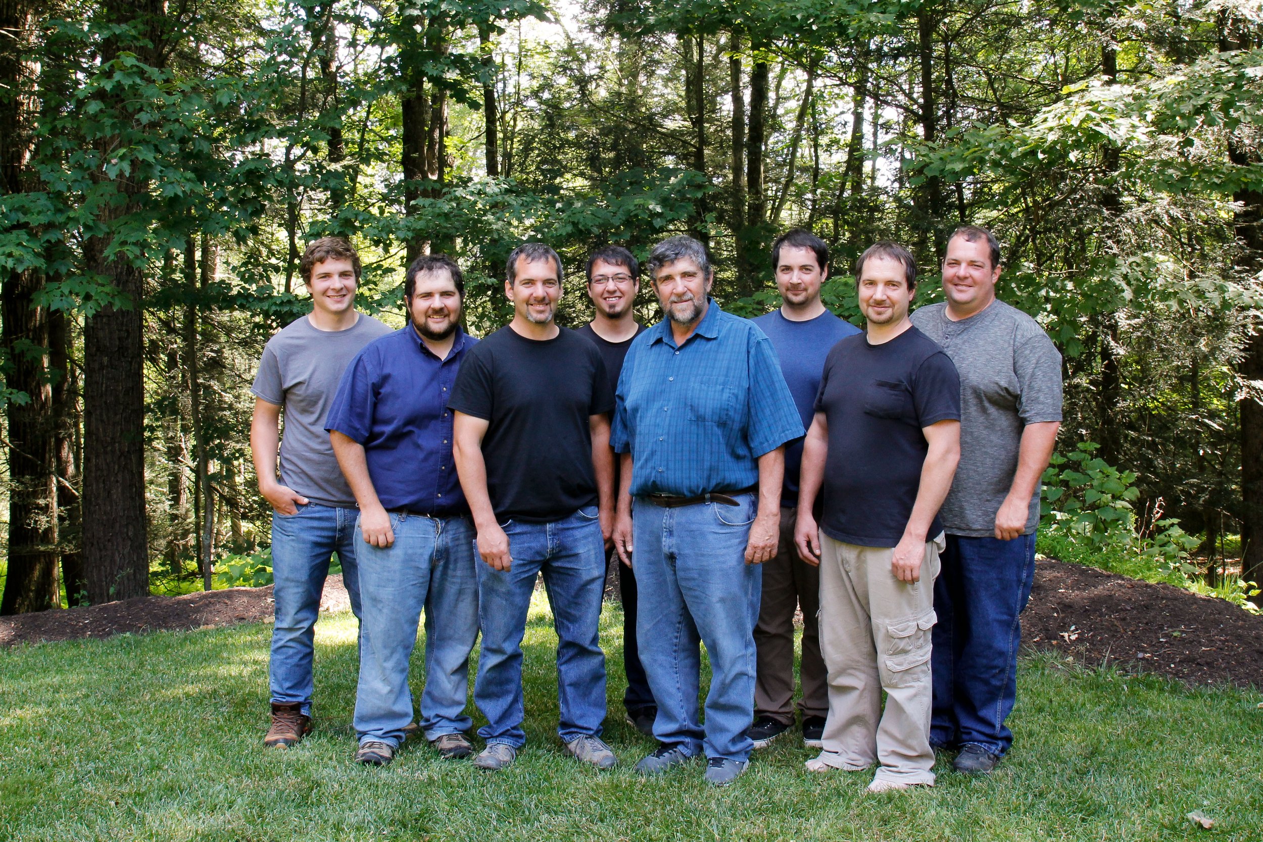 Group of eight men standing together in a forested area, smiling and dressed casually in outdoor clothing, surrounded by lush green trees and grass.